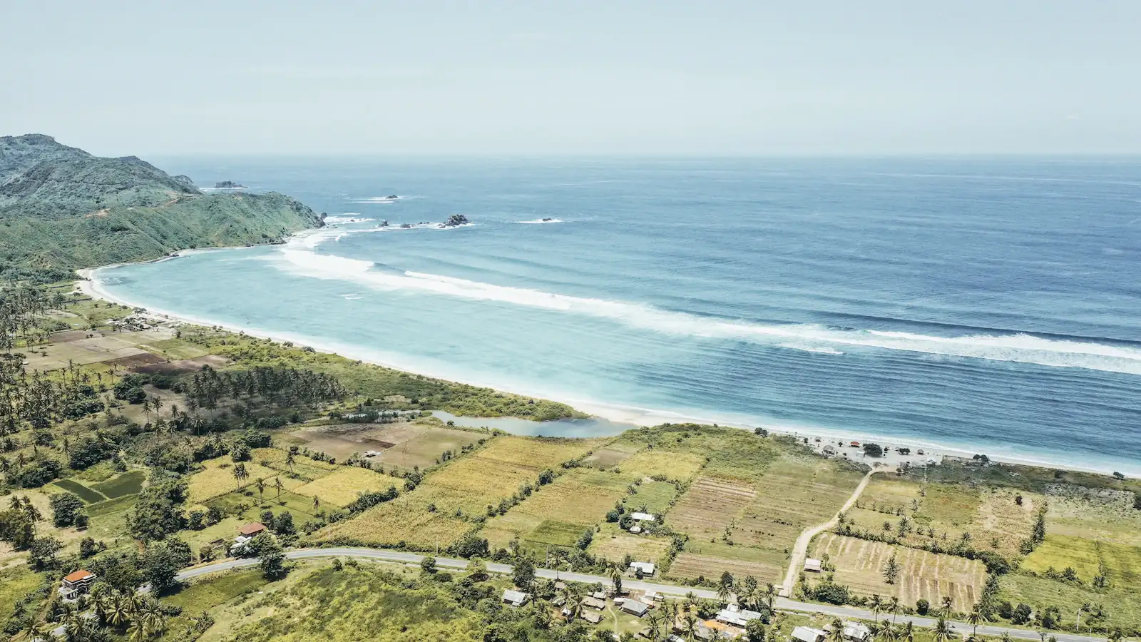 Aerial view of a south Lombok bay — surf lines along a white beach, palm groves and rice fields behind it
